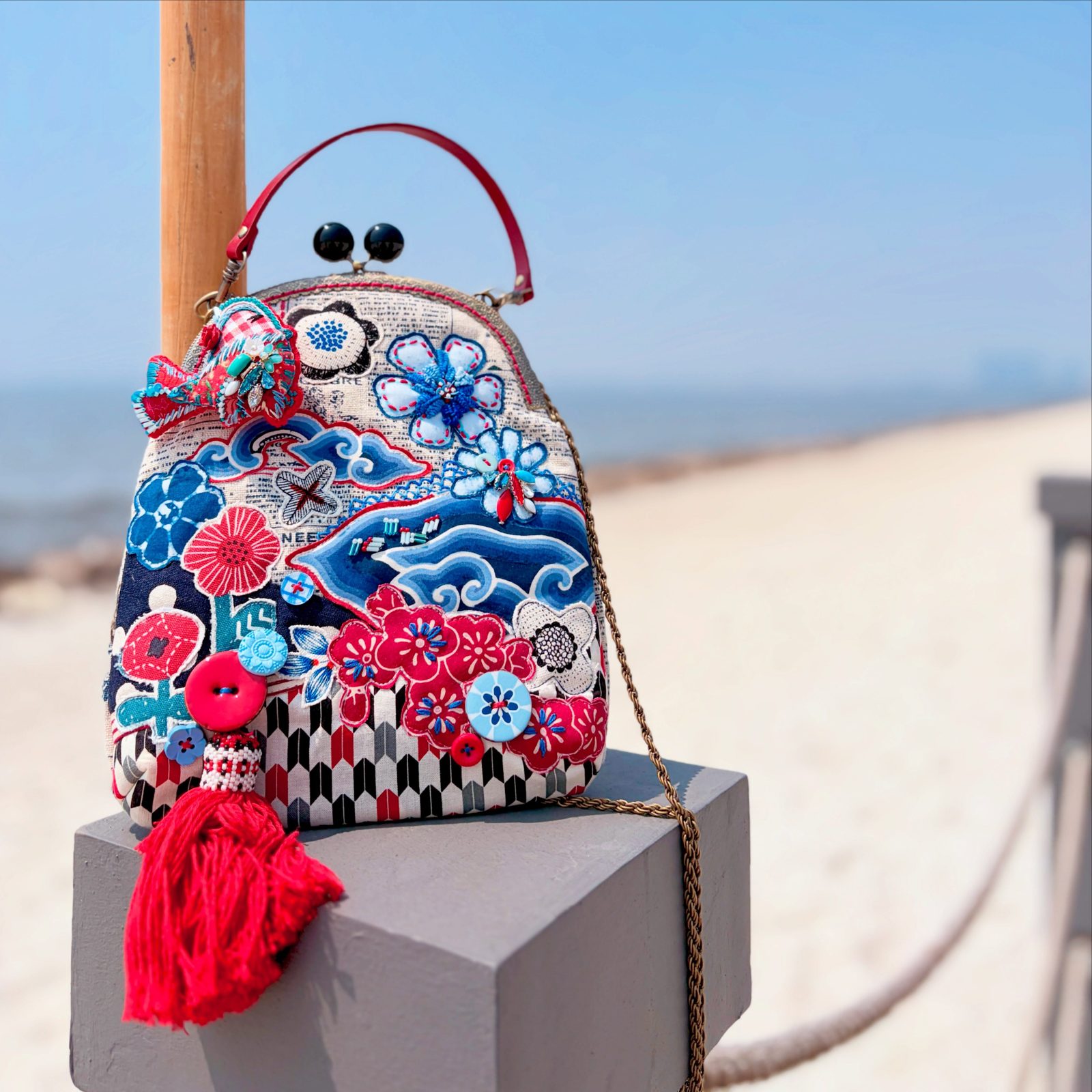 Batik floral kiss-lock bag with a red tassel, photographed on a bright beach