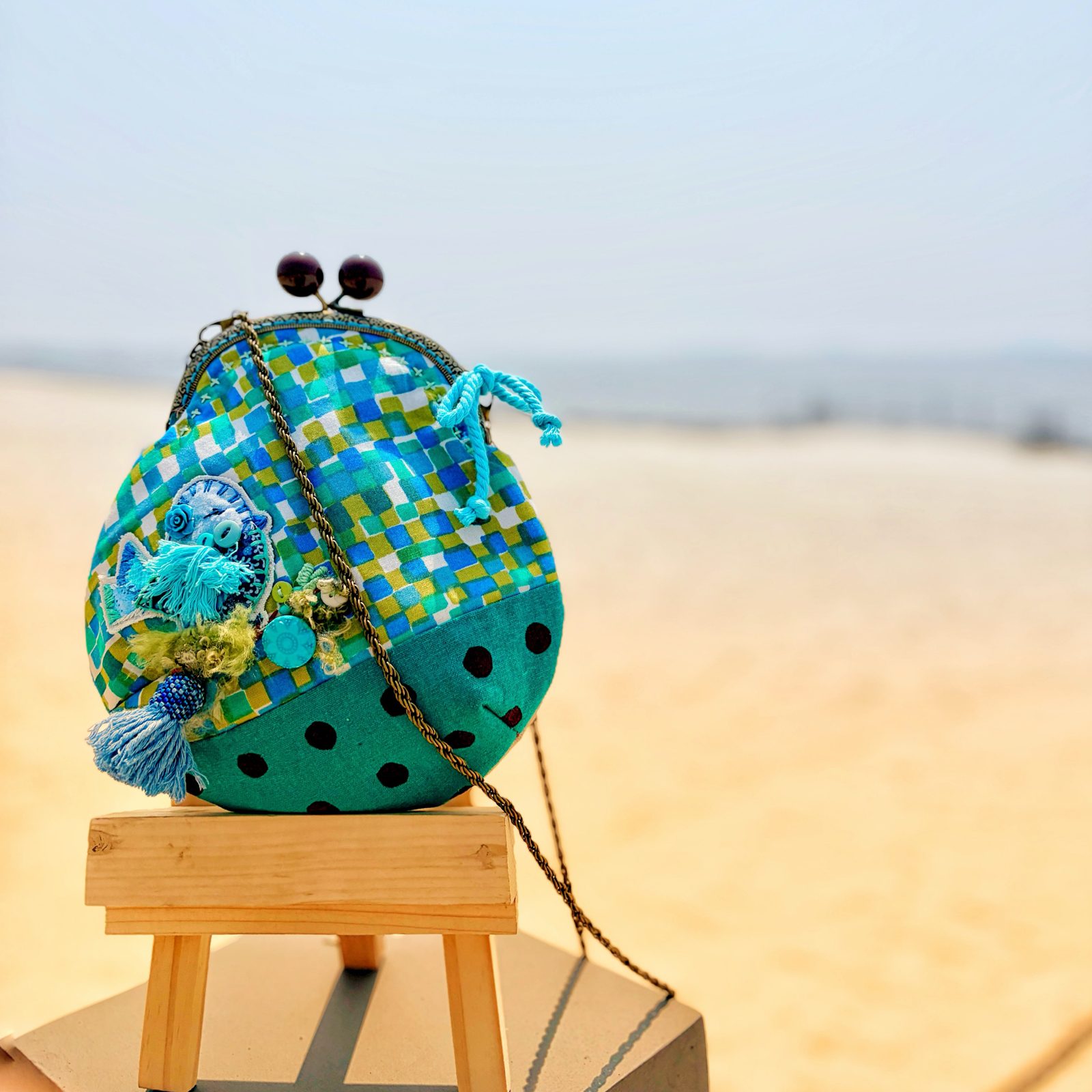 Teal polka-dot kiss-lock pouch with sequins and charms, on a wooden stool by the sea