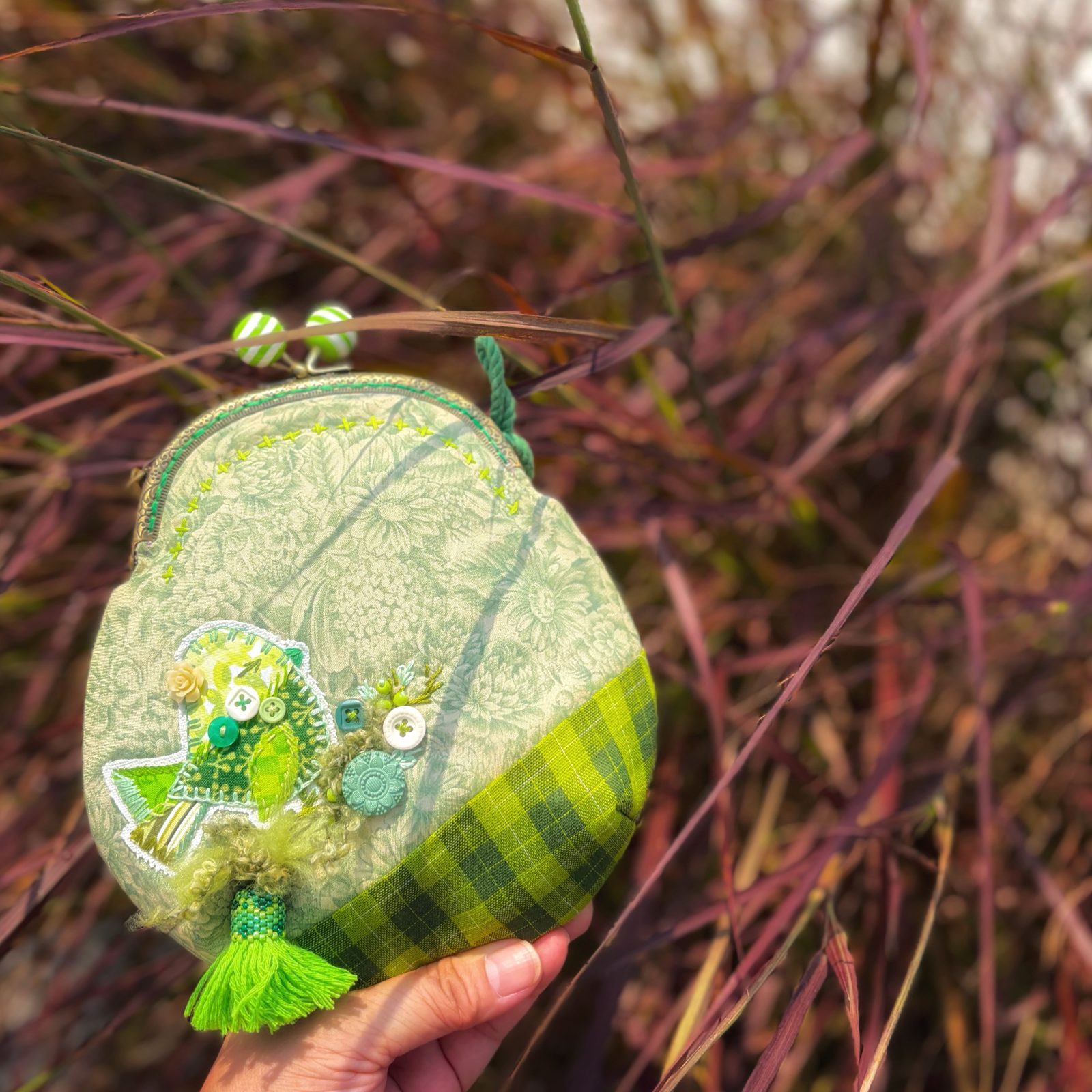 Pale green sling pouch with a frog charm and tassel, held up in tall grass